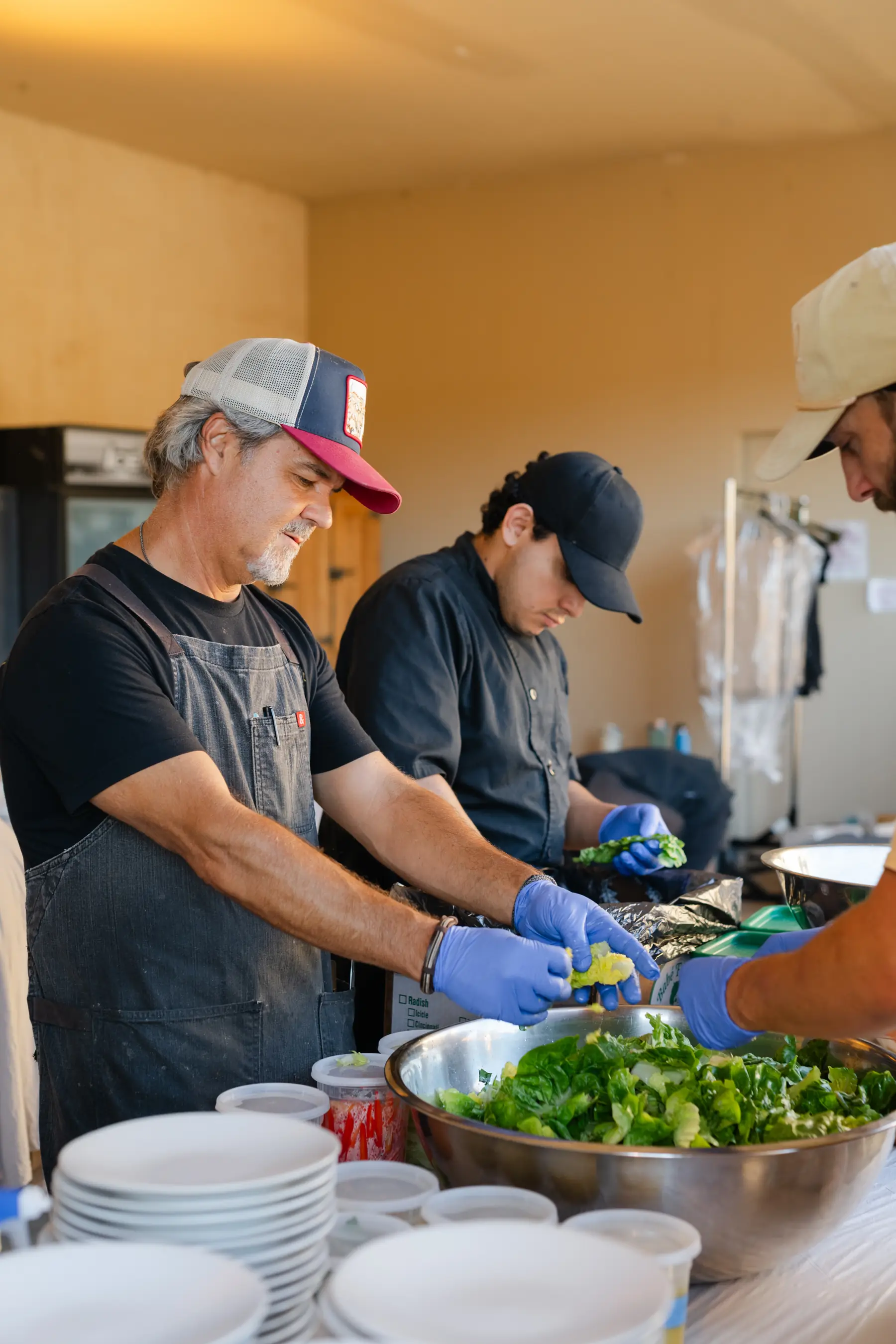 Three of the culinary team tearing salad greens into a mixing bowl behind stacks of plates