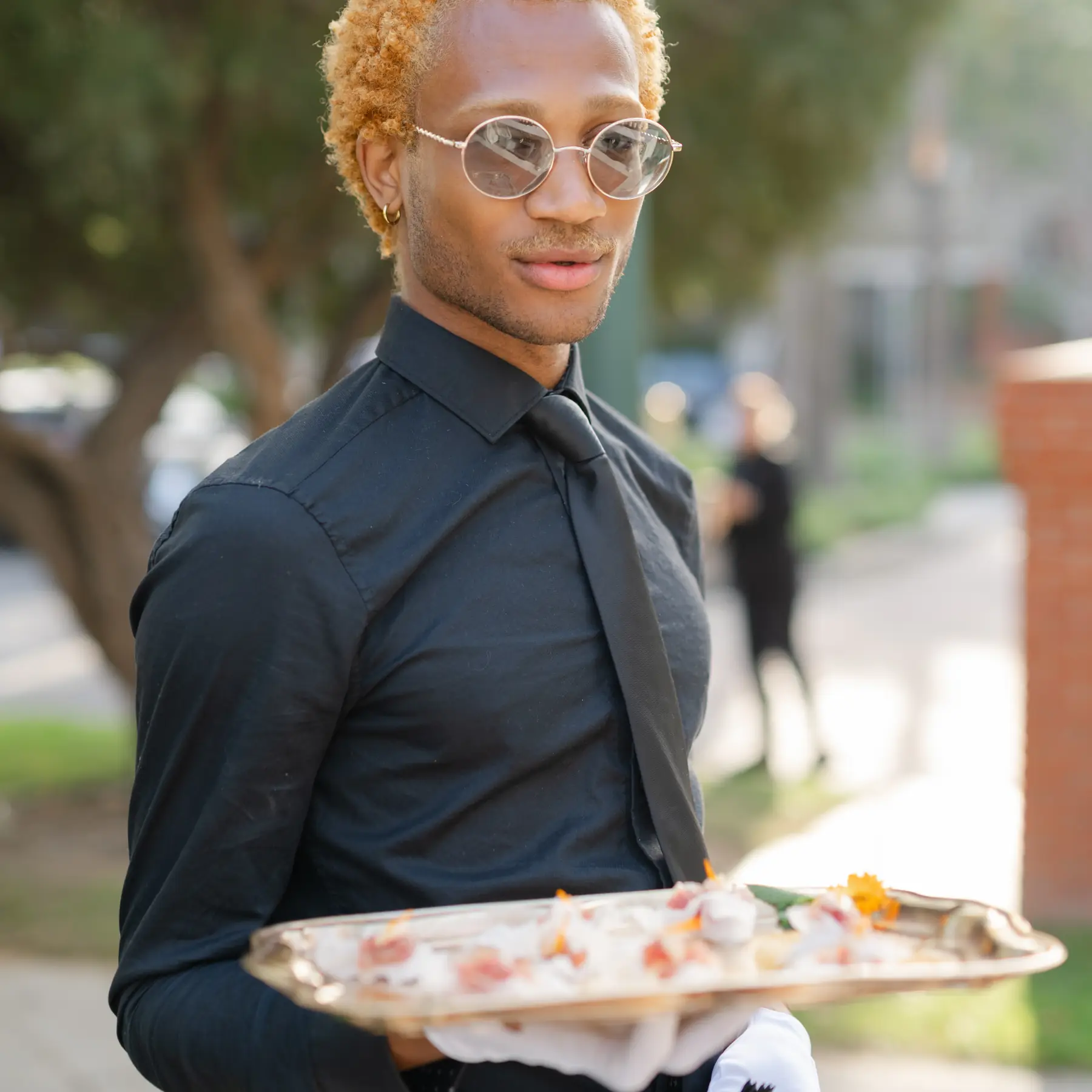 A server in a black shirt and tie carrying a silver tray of canapés outdoors