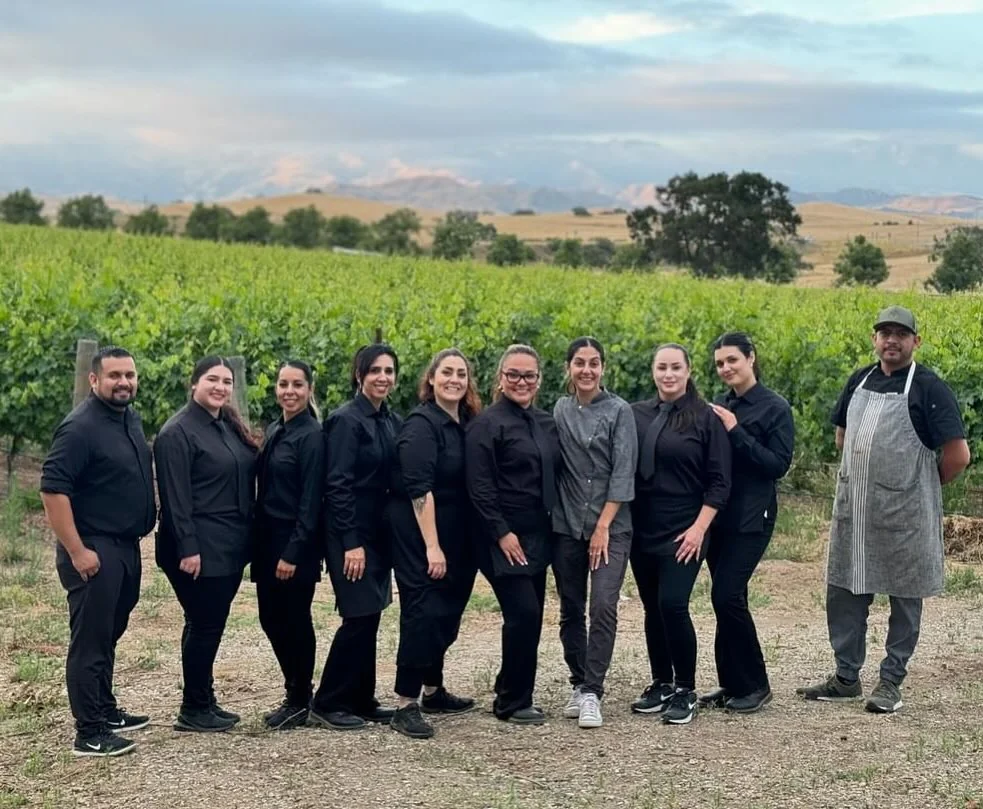 The All Purpose Flower culinary and service team standing together at the edge of a vineyard, with rolling hills and mountains behind them