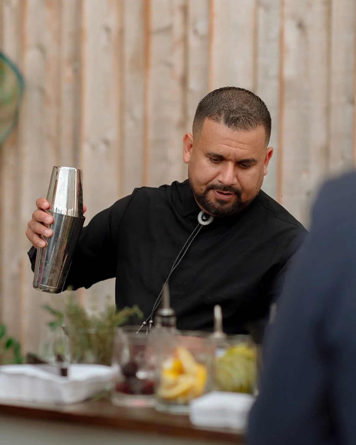 A bartender in black shaking a cocktail, with herbs, cherries and sliced fruit laid out along the bar