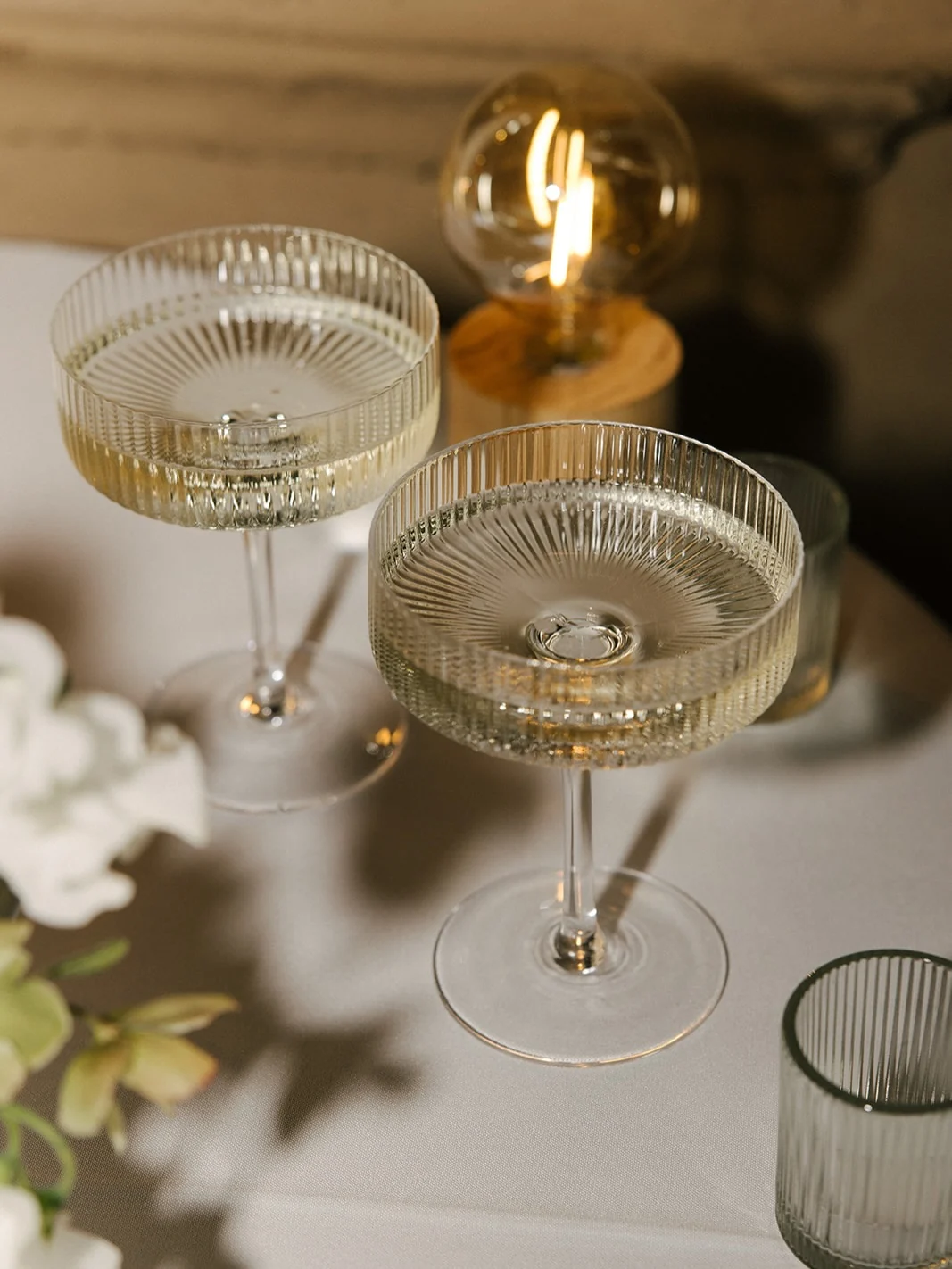 Two ribbed coupe glasses on a white table, with white flowers and a warm Edison bulb glowing behind