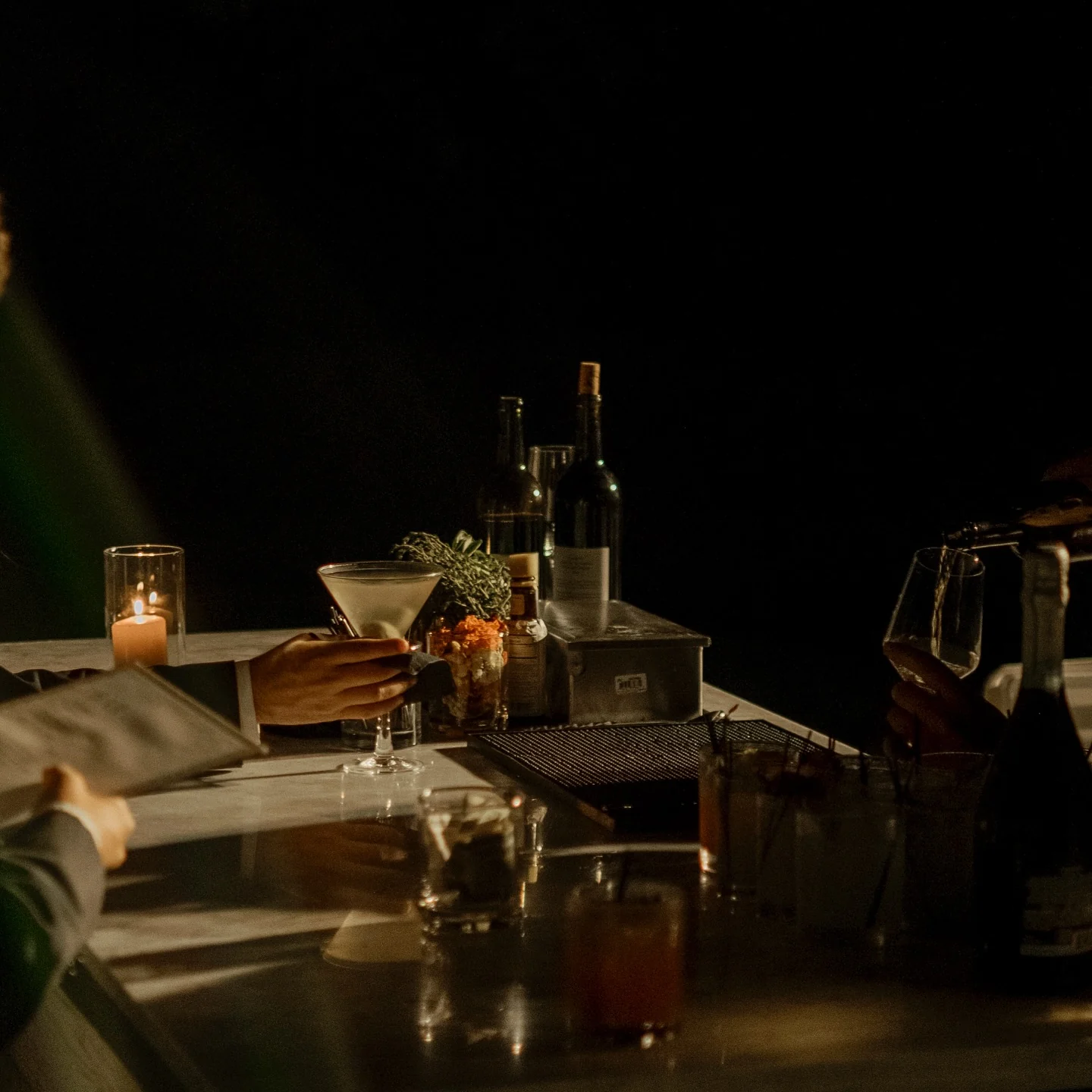 A candlelit bar after dark, a hand lifting a stemmed cocktail beside wine bottles and a lit votive