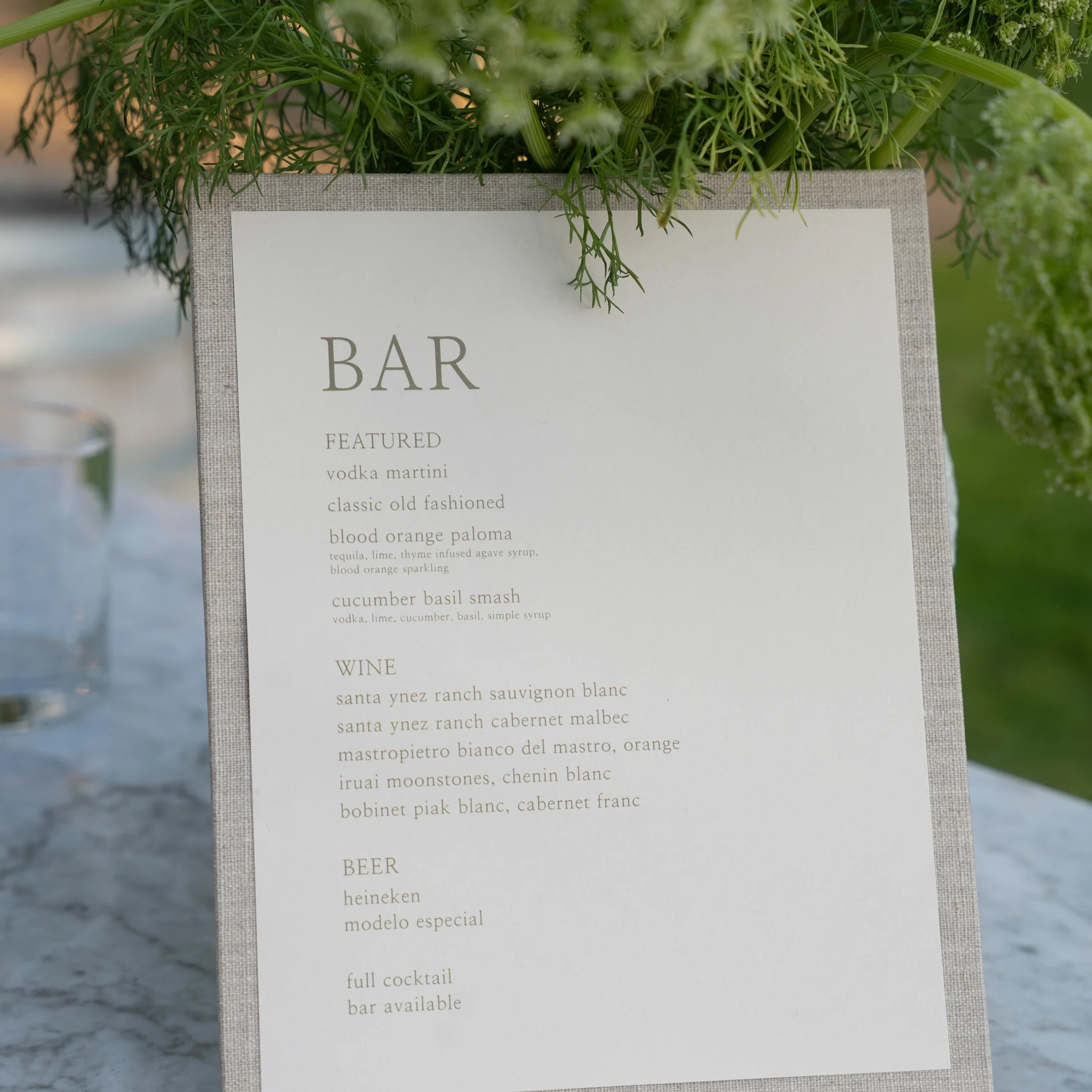 A printed bar menu card on a linen board, propped against greenery on a stone table