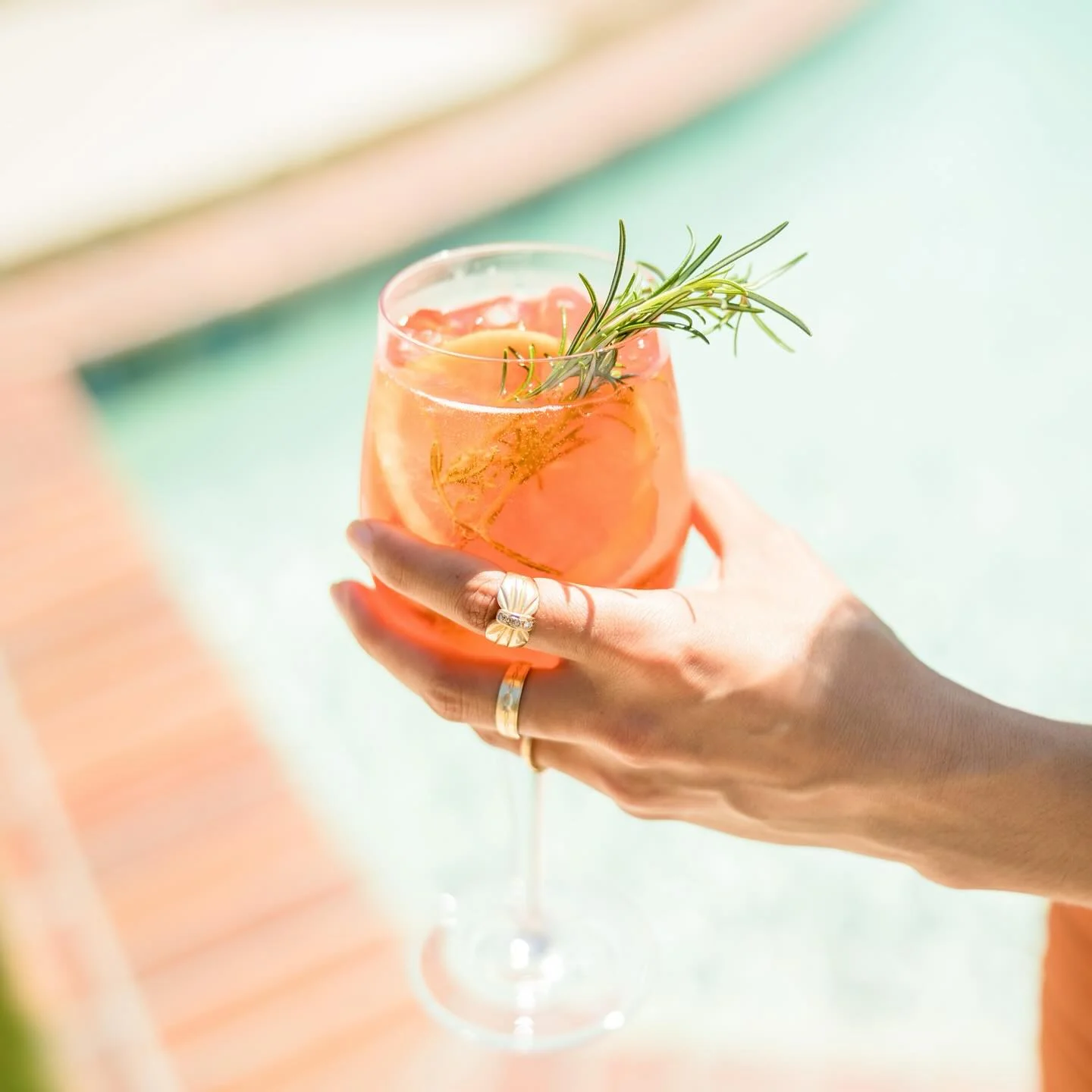 A hand holding a spritz garnished with a rosemary sprig above bright turquoise pool water