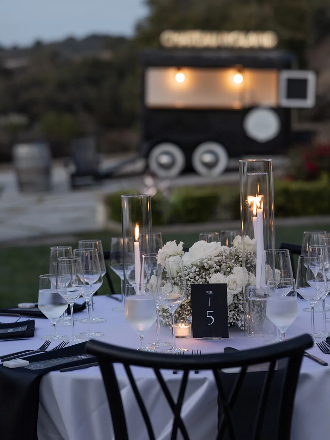 A round table set with candles and white florals in front of the lit Chateau Noland bar trailer