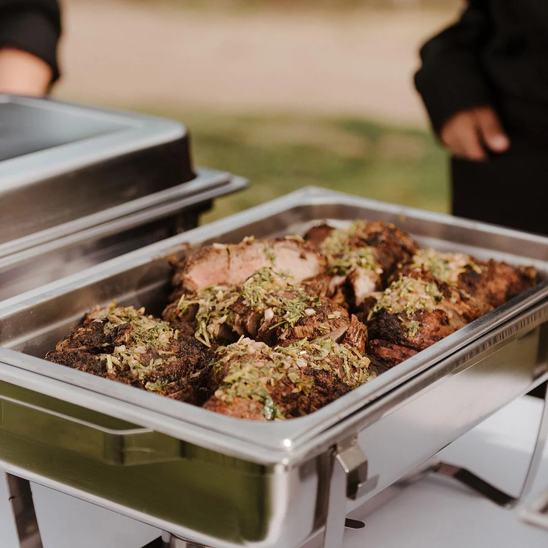 A chafing pan of sliced roast meat finished with a fresh herb and shallot relish on an outdoor service line