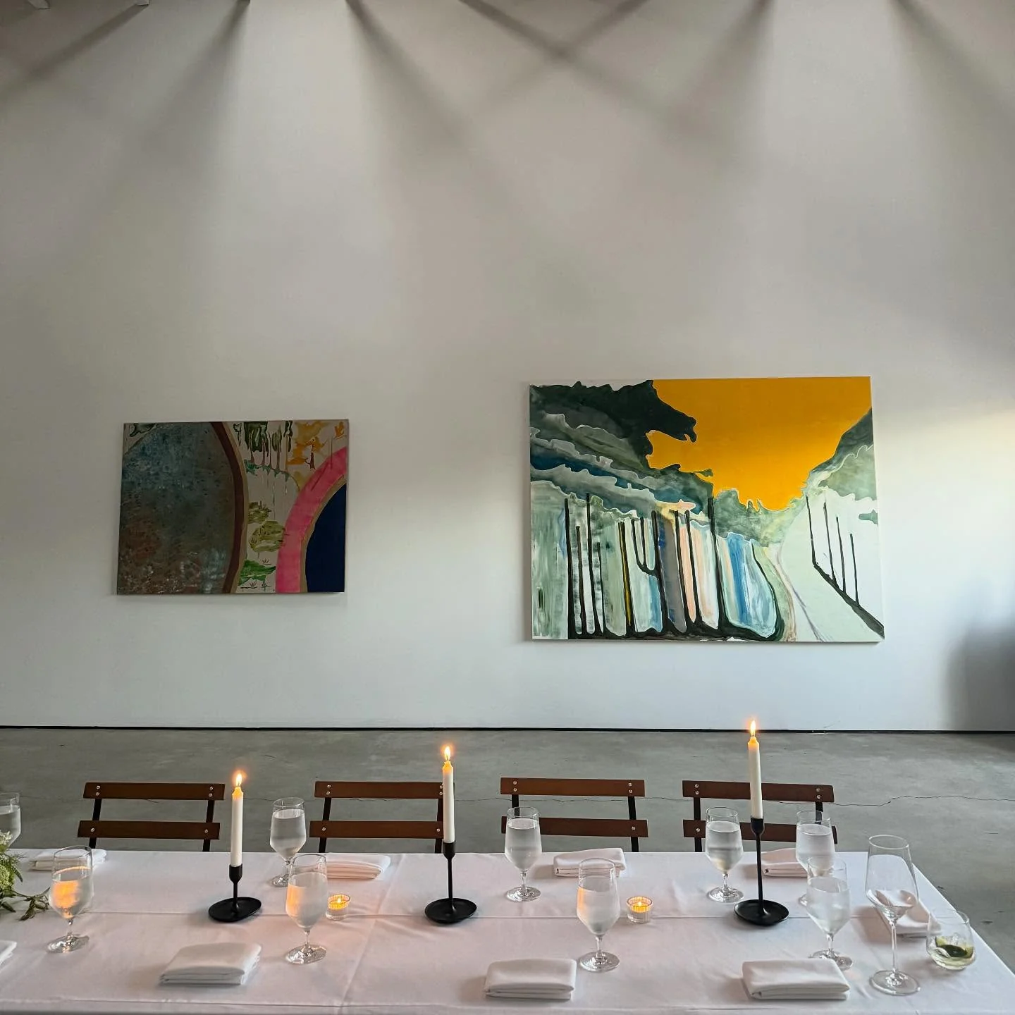 A long table set with white linen, taper candles and glassware in a white-walled room hung with two large artworks