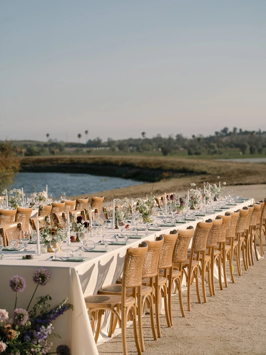 A long table beside the pond at Gundlach Bundschu, set with ivory linen, sage napkins, white taper candles and low white and burgundy florals, lined with cane-backed chairs