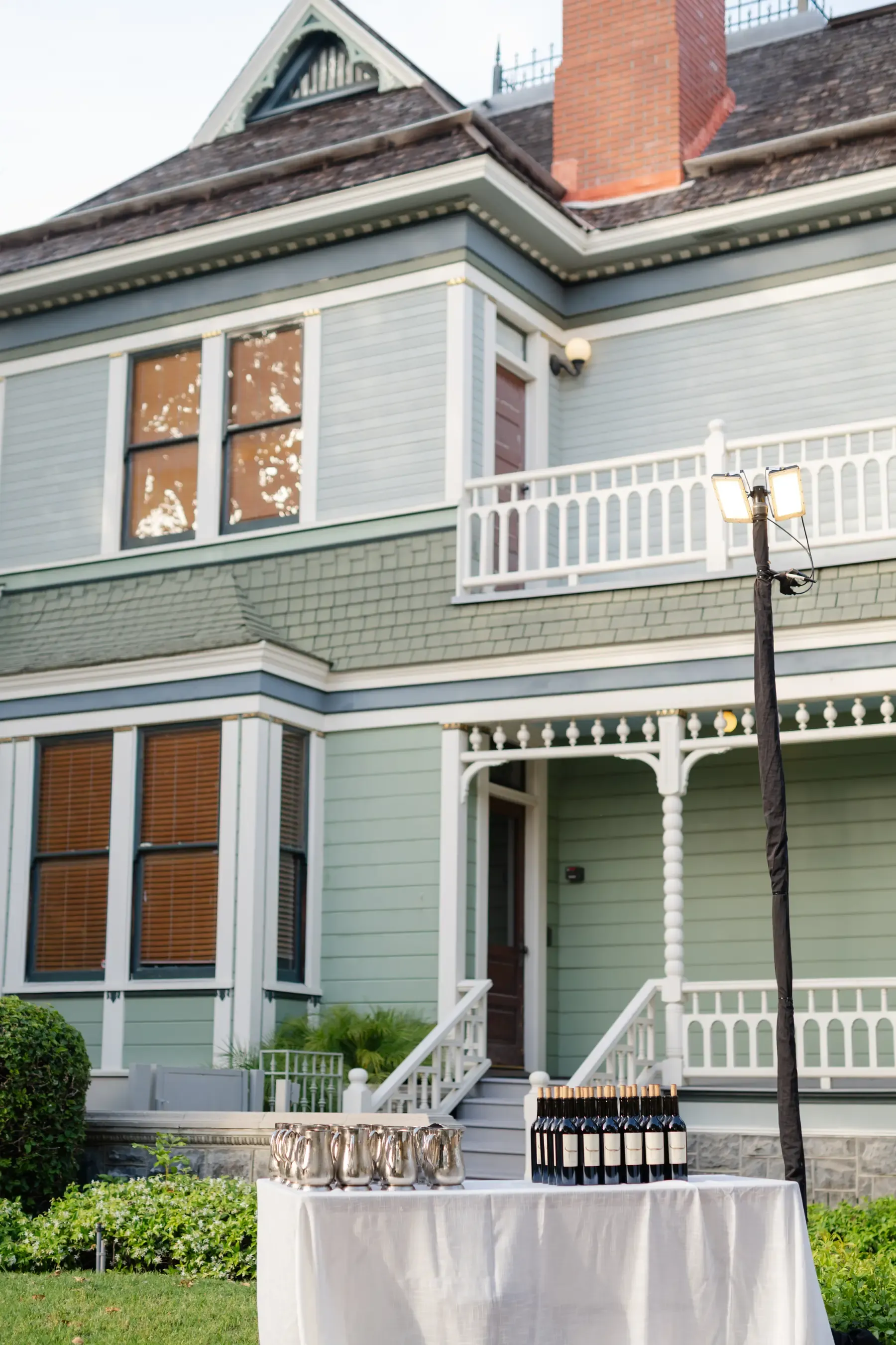 A sage-green Victorian with a spindled porch at Heritage Square, silver pitchers and a rank of wine bottles set out on a linen-draped table on the lawn below it