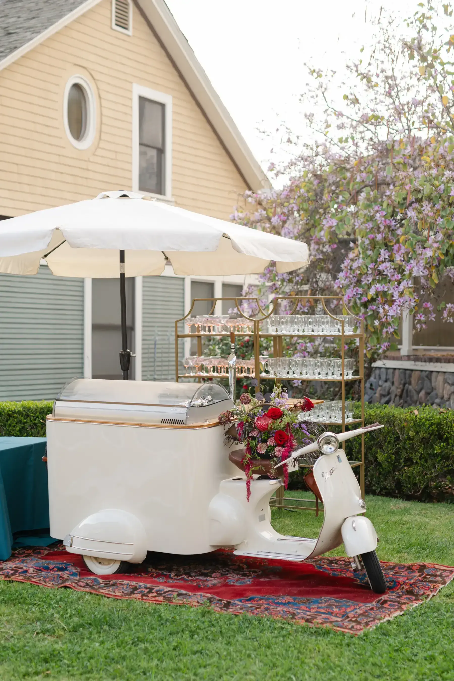 A vintage white Vespa bar cart under a cream umbrella on a Persian rug at Heritage Square, a gold étagère of glassware and deep red florals beside it