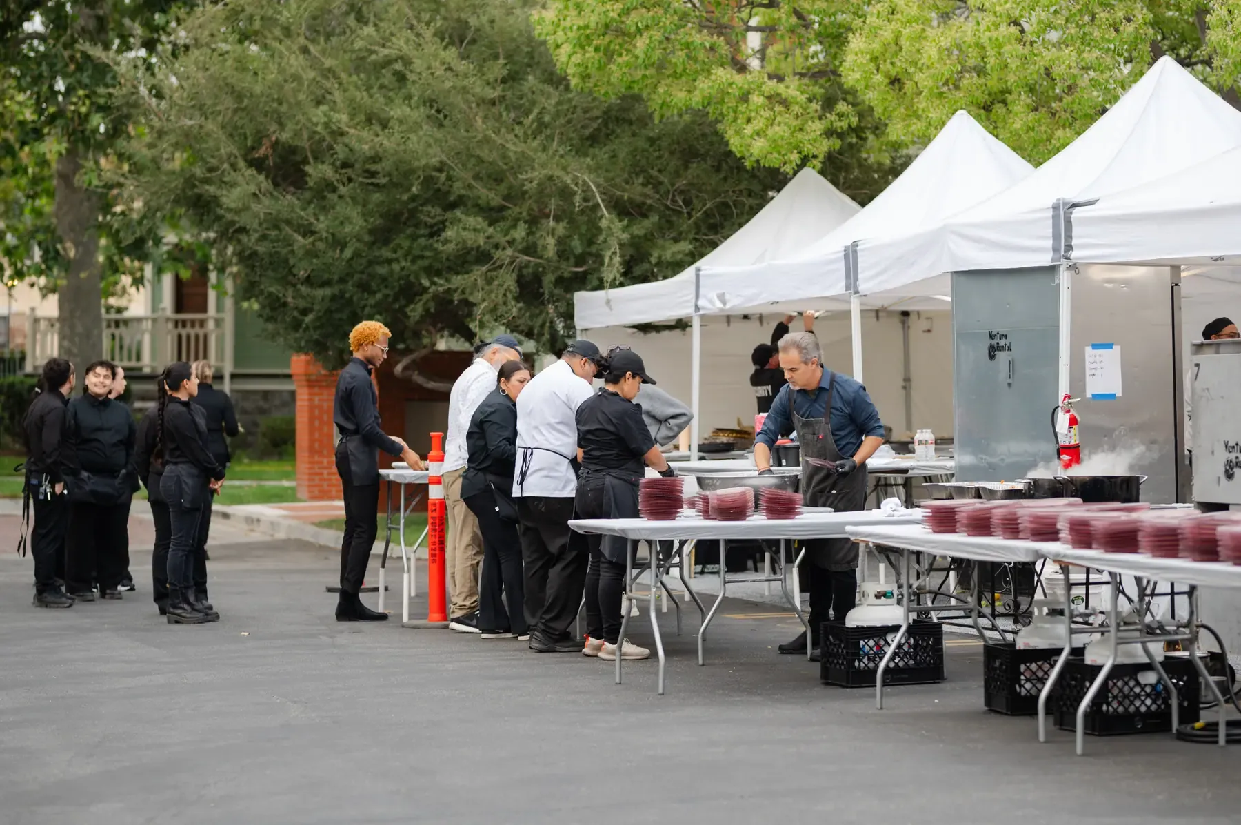 The tented satellite kitchen we set up at Heritage Square — white canopies on the pavement, stacked plates and hot boxes along folding tables, the culinary team on the line and the service team waiting to run