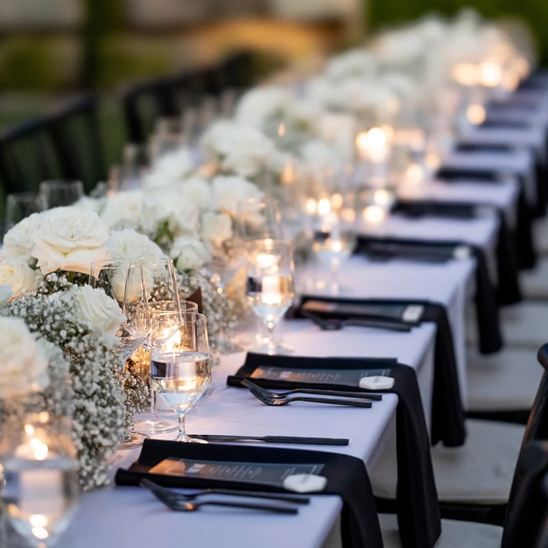 A long table lined with white roses and candlelight, set with black napkins and printed menus