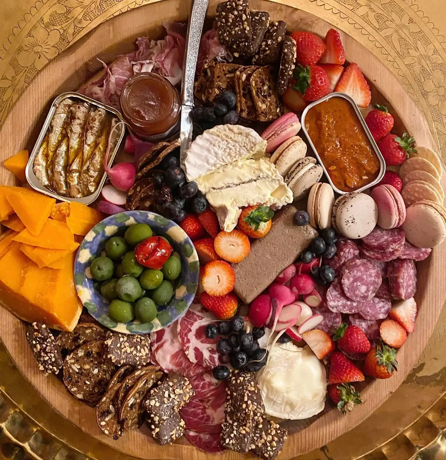 An overhead grazing board on a round wooden platter set on an engraved brass tray — a cut wheel of brie, pâté, sliced salami and prosciutto, wedges of aged cheddar, a tin of sardines, a bowl of green olives, strawberries, blueberries, seeded crackers and pastel macarons