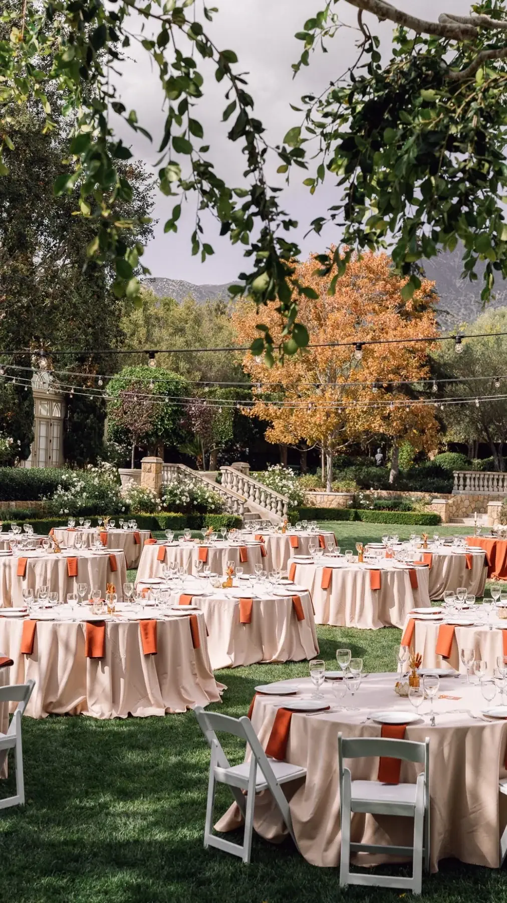 Round tables in ivory linen with coral napkins, set on a lawn beneath a spreading oak
