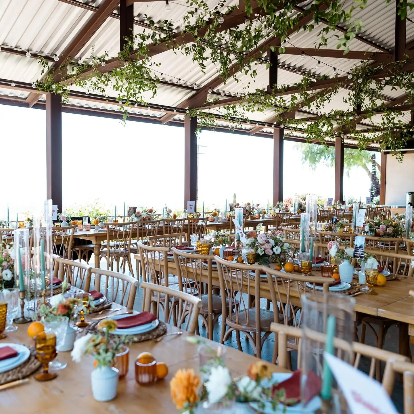 Long banquet tables under an open-sided pavilion hung with trailing greenery and string lights