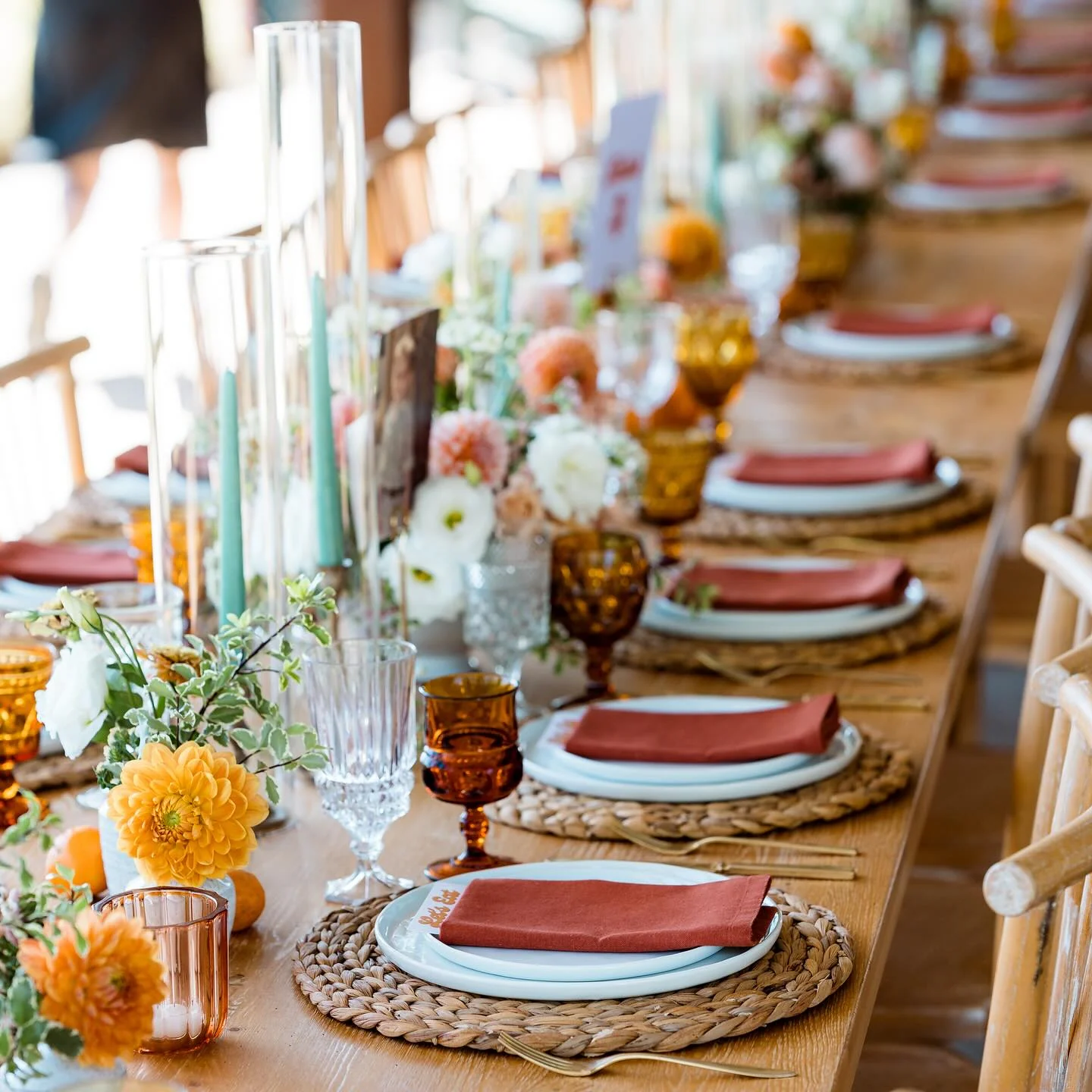A long wood table set with rust napkins, woven chargers, amber glassware and low floral arrangements