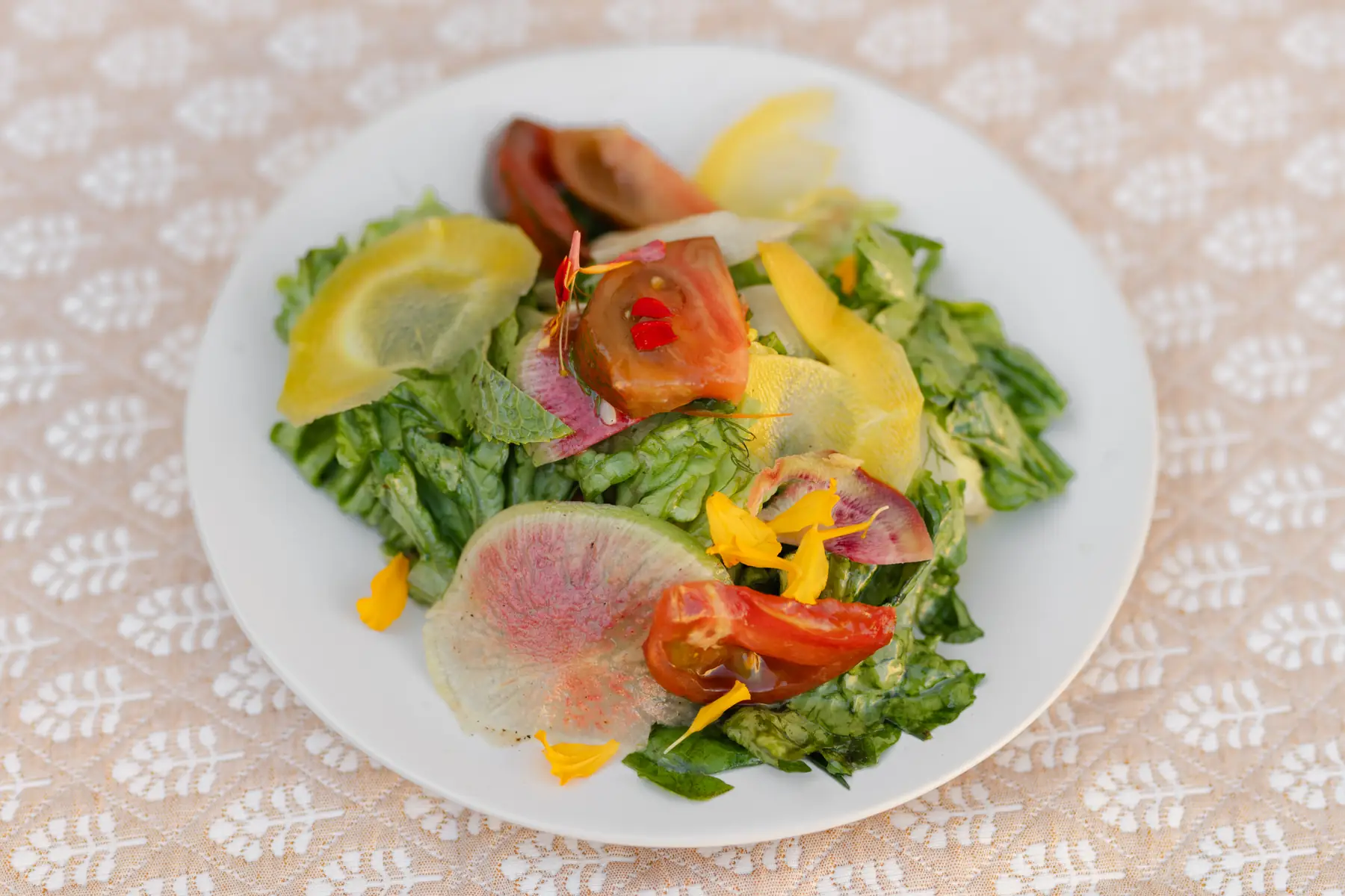 A plated salad of little gem leaves with tomato, shaved watermelon radish and squash ribbons, finished with edible flower petals