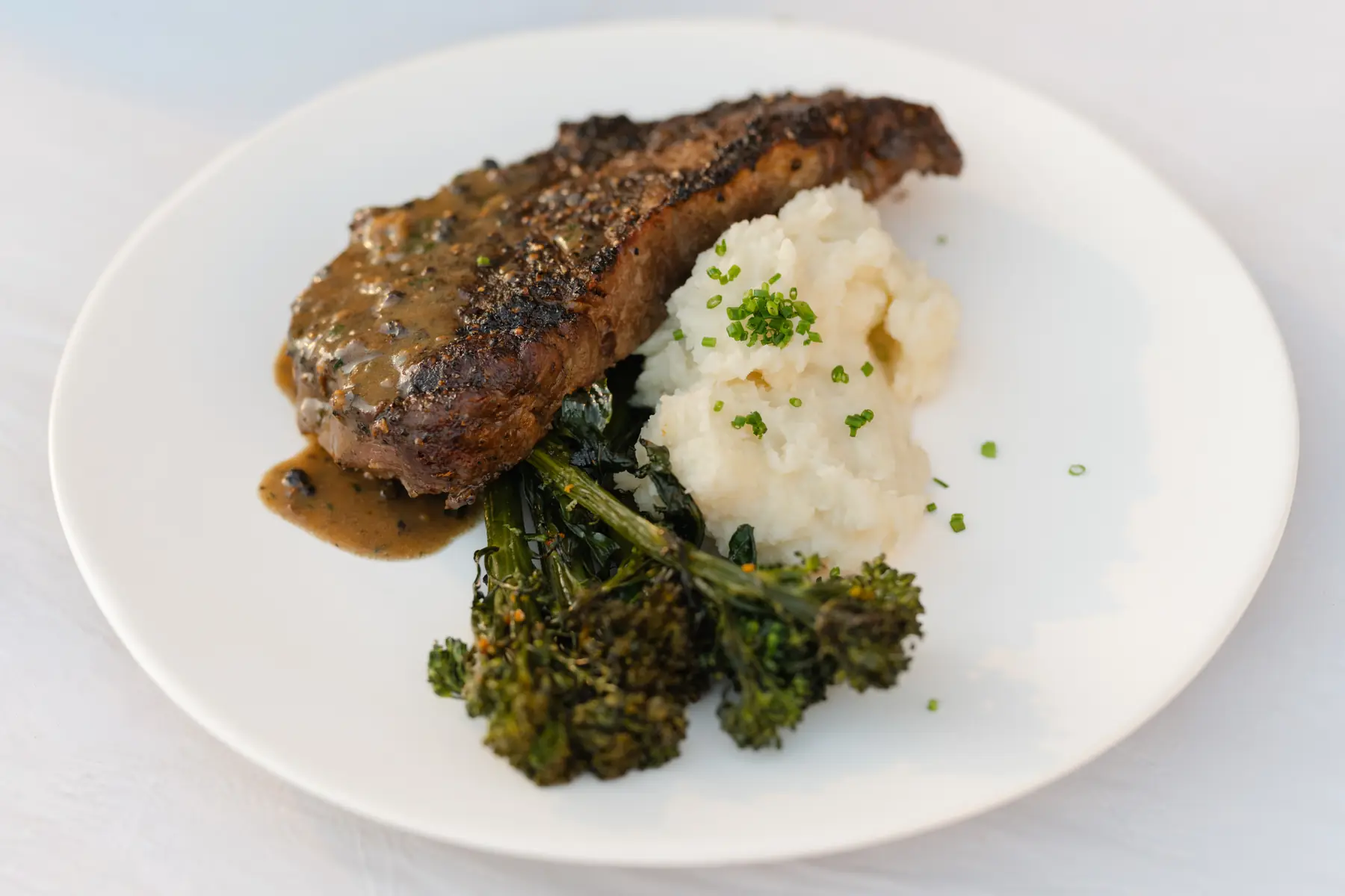 A plated New York steak au poivre with potato purée and roasted broccolini