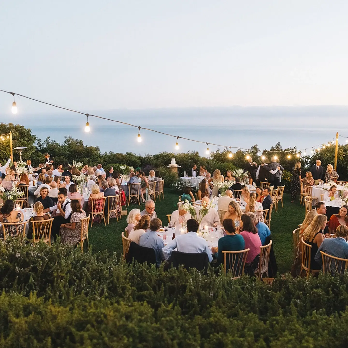 Guests at round tables under strung lights at dusk at Regina Ranch, the ocean beyond the hedge