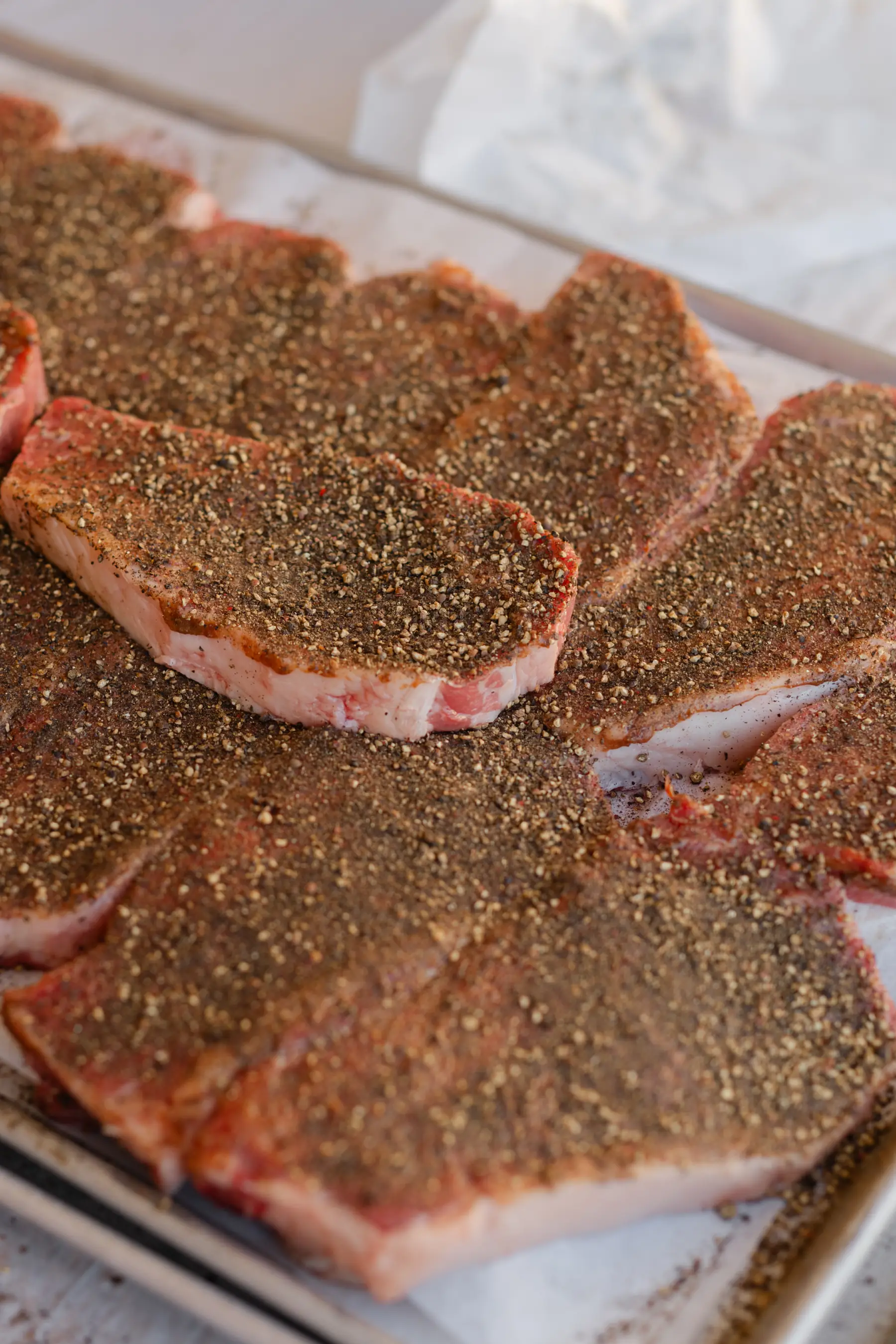 Steaks crusted in cracked pepper on a sheet tray, ready for the grill