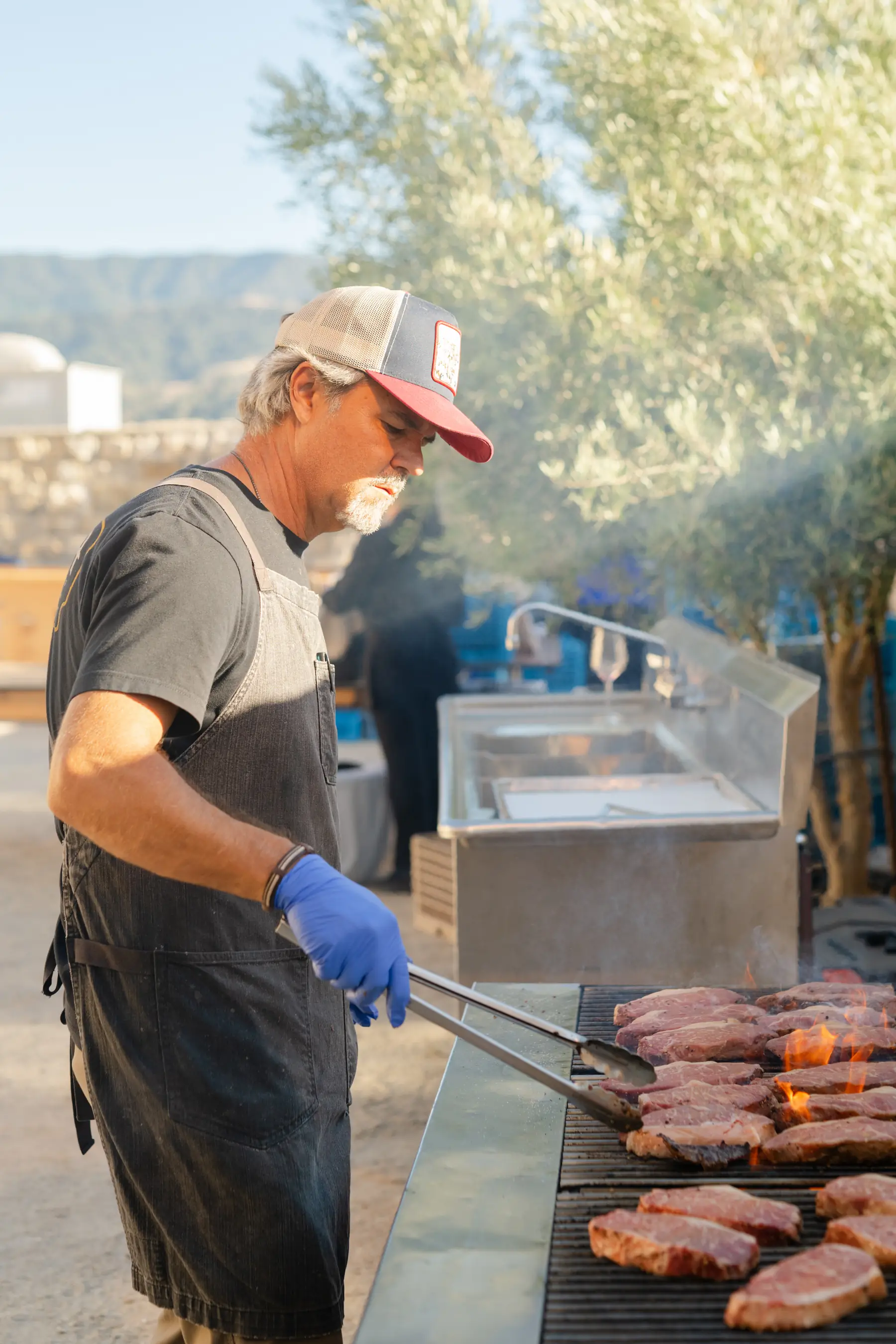 A chef tending steaks over open flame on the grill line under the olive trees at Sunstone Villa, hills behind