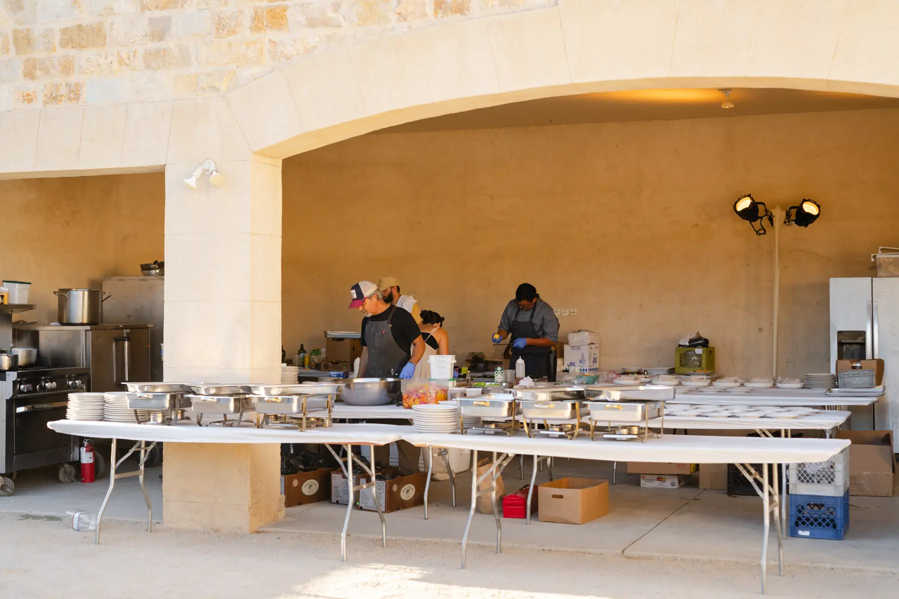 The field kitchen and catering line built under the stone arcade at Sunstone Villa, chafing dishes and stacked plates along the long tables