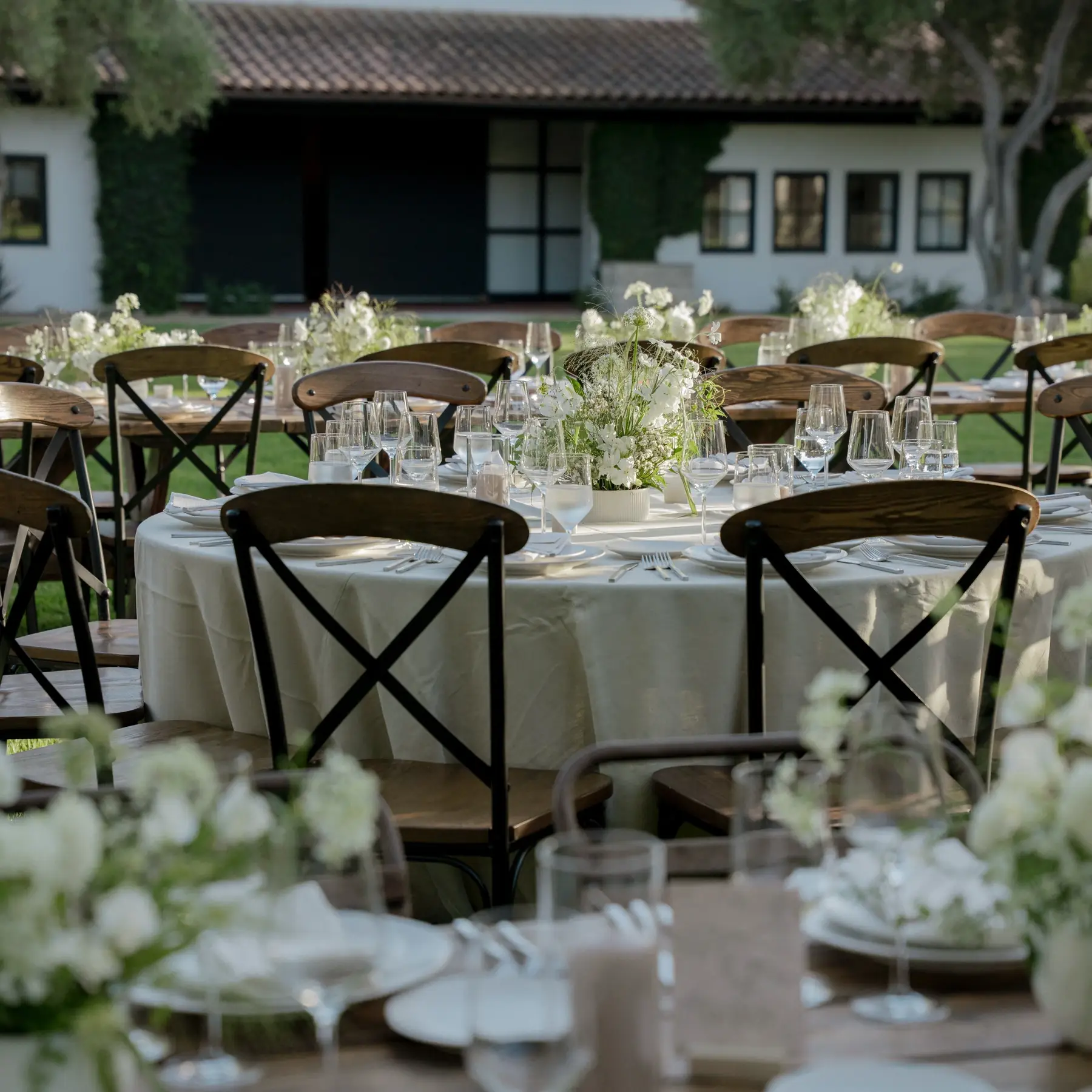Round tables in ivory linen with white florals on the lawn at Santa Ynez Ranch, the white tile-roofed estate behind