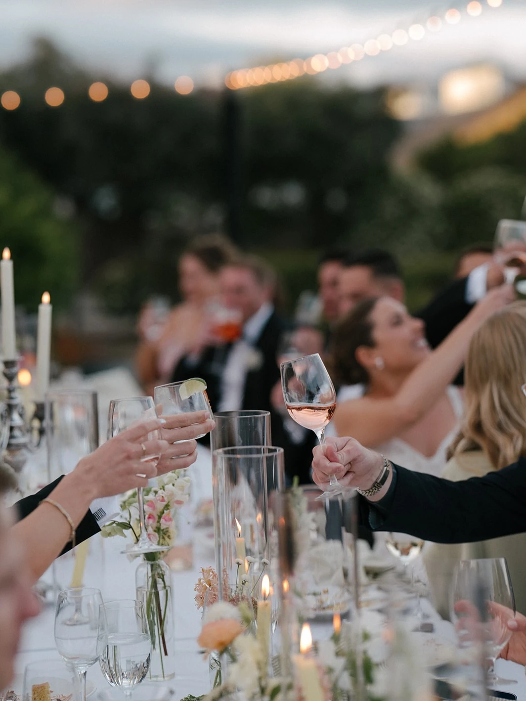 A toast down a candlelit long table at dusk, glasses raised over blush and white florals