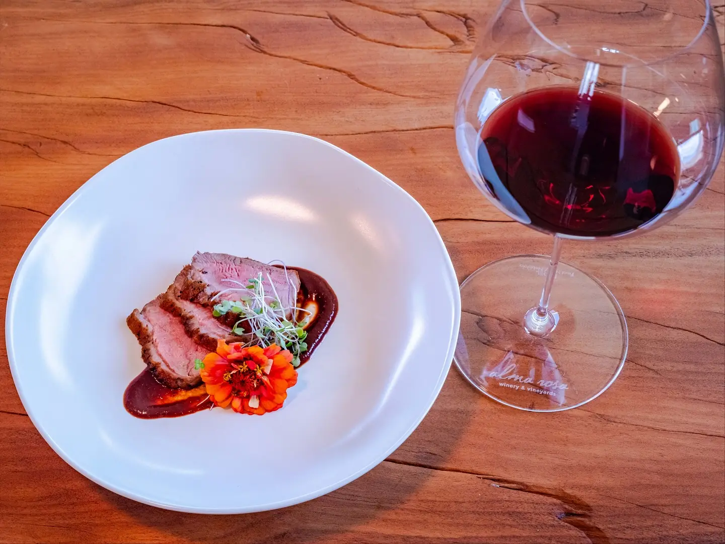 Sliced duck breast on a dark cherry and rosemary jus with microgreens and an orange zinnia, beside a glass of Pinot Noir etched with the Alma Rosa winery mark