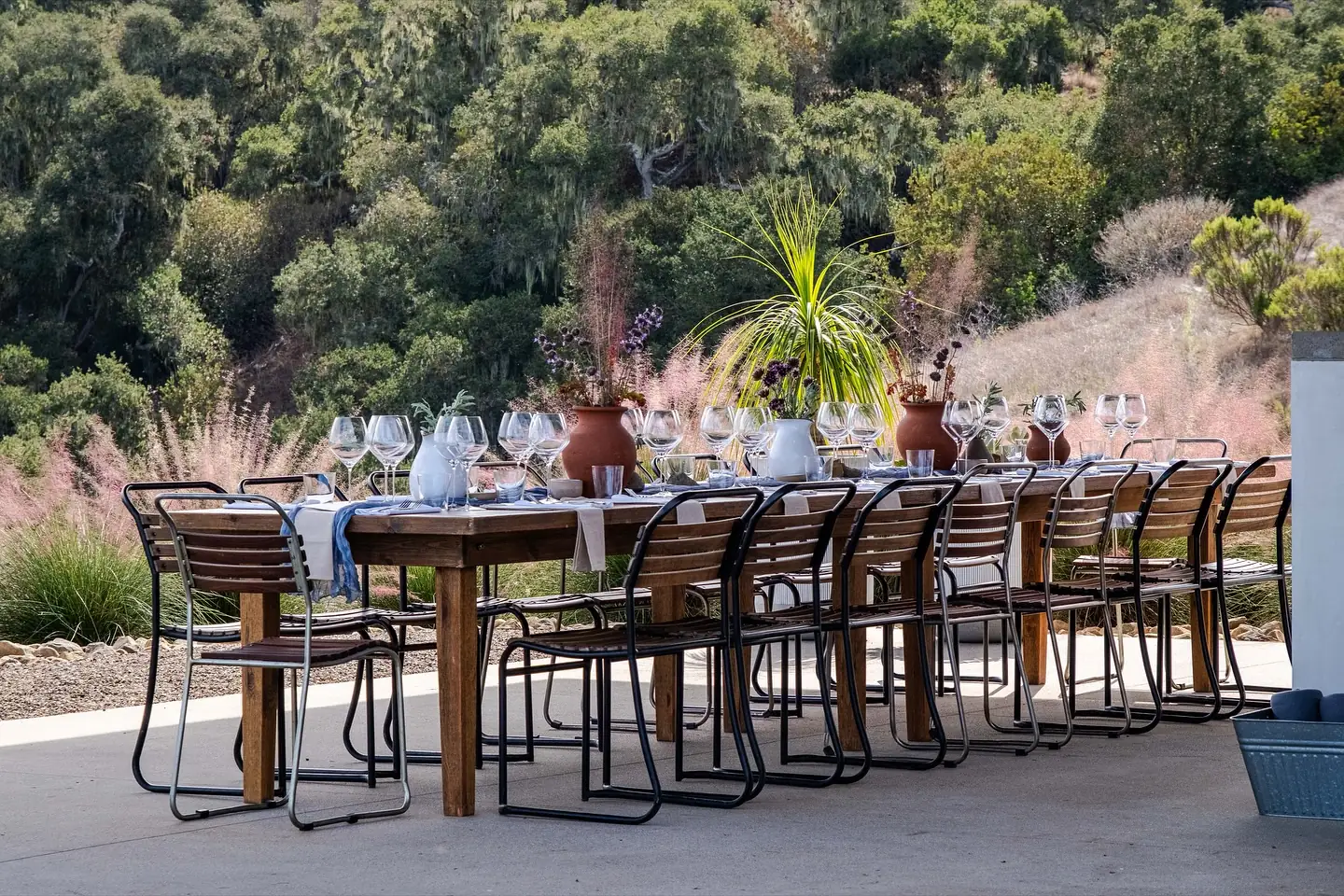 A long wooden table laid with wine glasses at every setting and terracotta pots of dried florals, ringed by black metal chairs on a concrete terrace above pink muhly grass and an oak hillside
