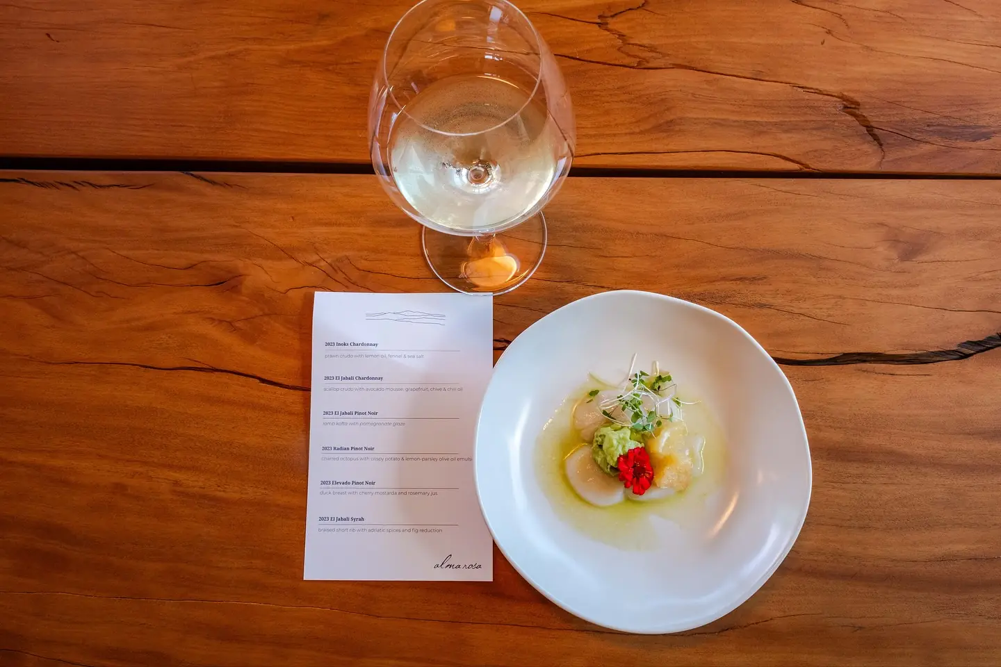 A printed pairing card listing six Alma Rosa wines against six courses, beside a plated scallop crudo with avocado mousse, grapefruit and a red flower, and a glass of white wine