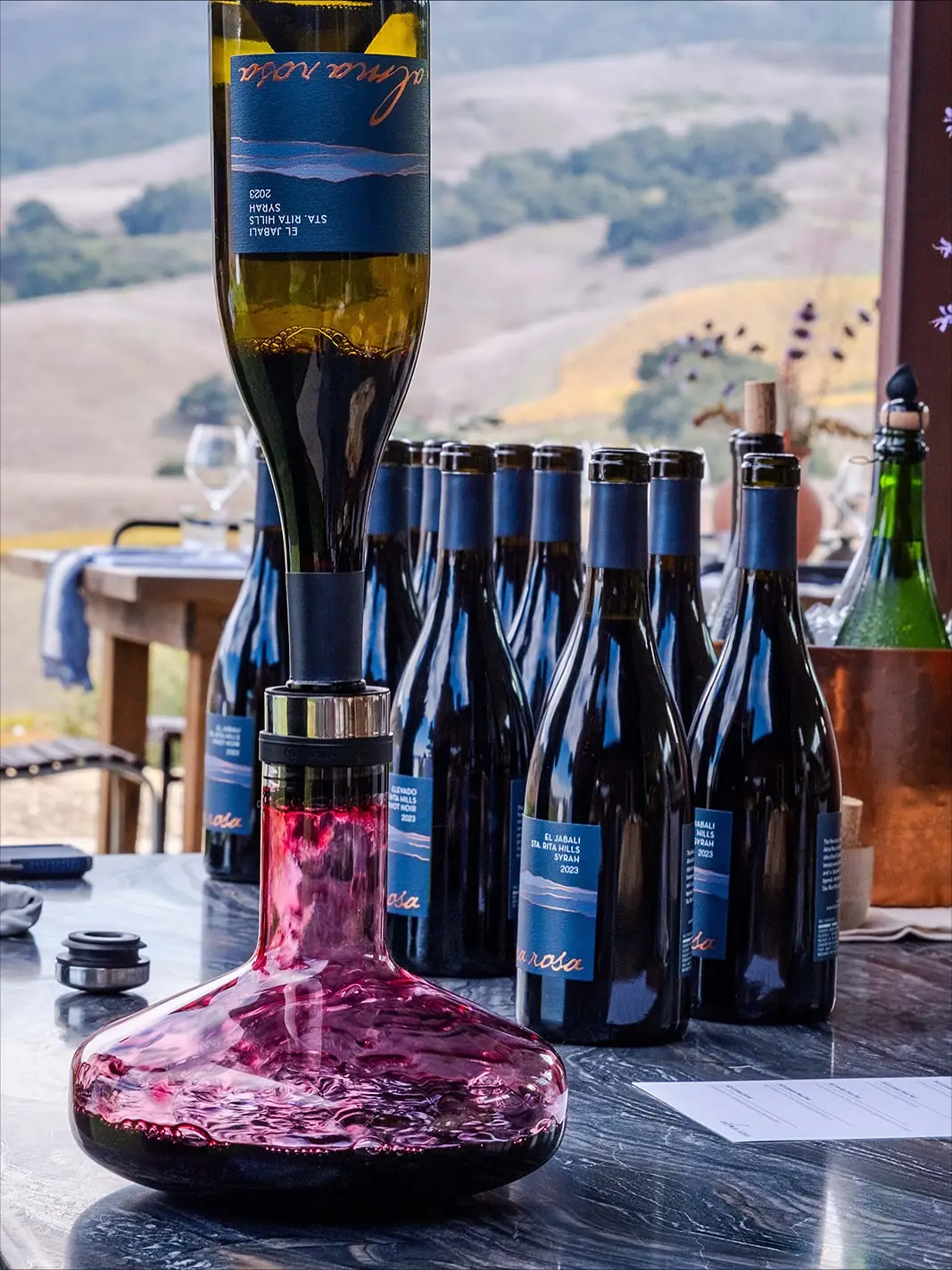A rank of Alma Rosa Sta. Rita Hills bottles beside a decanter of red wine on a marble bar, an upended Syrah bottle in a pouring cradle above, vineyard hills behind